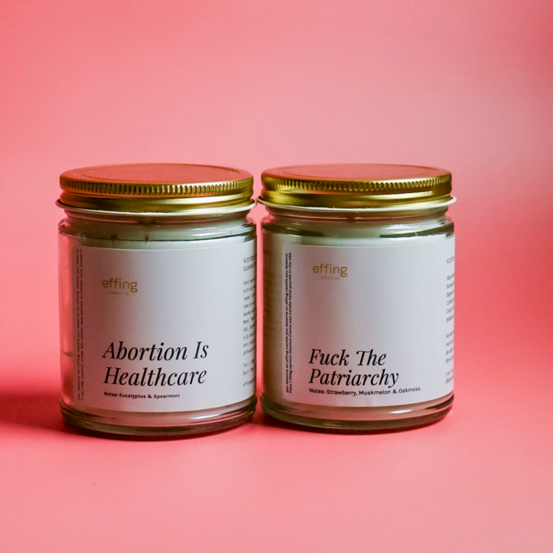 Abortion is healthcare candle next to a Fuck the Patriarchy candle. They are on a pink background.