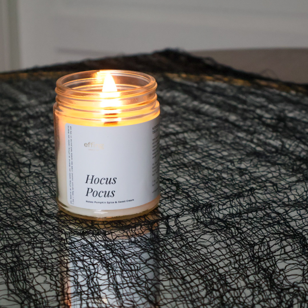 A lit candle in a clear glass jar with a white label reading 'Hocus Pocus' placed on a black mesh fabric surface.