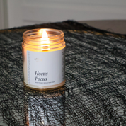 A lit candle in a clear glass jar with a white label reading 'Hocus Pocus' placed on a black mesh fabric surface.