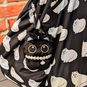 A close-up of a black reusable bag with a white cat print, featuring a cute black plush cat charm with embroidered eyes, a pearl necklace, and a small silver bell.