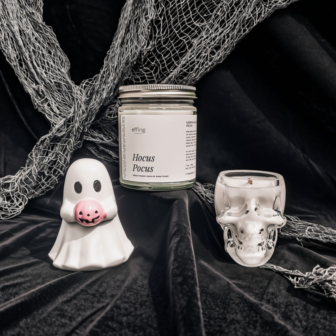 A styled product shot featuring a 'Hocus Pocus' candle by the brand effing, displayed alongside a white ghost figurine holding a pink pumpkin and a clear glass skull candle holder, all set against a black velvet background with draped netting.
