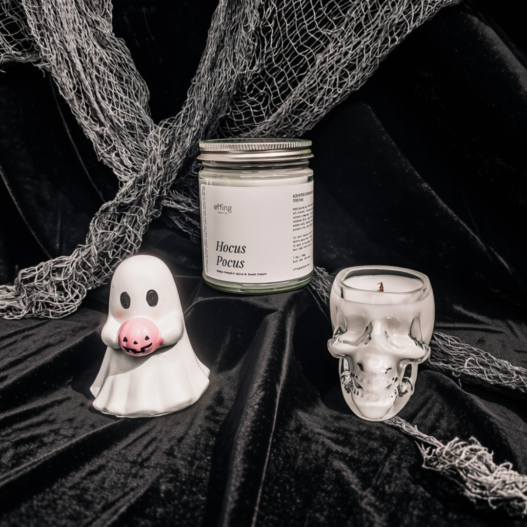 A styled product display featuring a white ghost figurine holding a pink pumpkin, a glass jar candle labeled 'Hocus Poems' by the brand 'etting', and a skull-shaped glass candle holder, all arranged on black velvet fabric with netting.