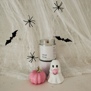 A Halloween-themed candle in a clear glass jar with a white label reading 'Hocus Pocus', styled with a small pink pumpkin and a white ghost figurine in front of a draped white fabric backdrop with black spider and bat cutouts.