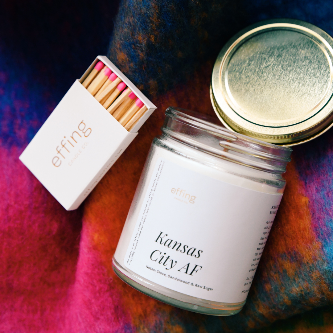 A glass jar candle with a white label that reads 'Kansas City AF', alongside a box of matches with pink tips, all resting on a colorful textured surface.