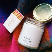 A glass jar candle with a white label that reads 'Kansas City AF', alongside a box of matches with pink tips, all resting on a colorful textured surface.