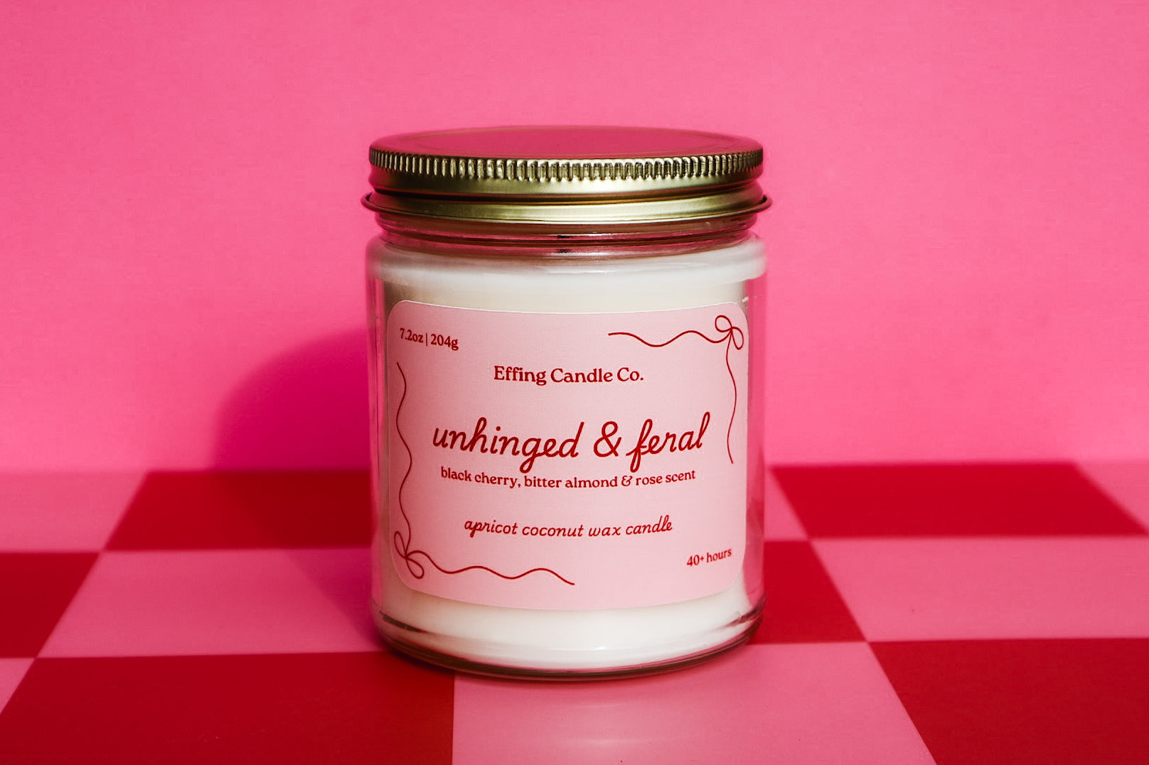 Candle jar that says Unhinged and Feral with a pink label on a red and pink checkered background