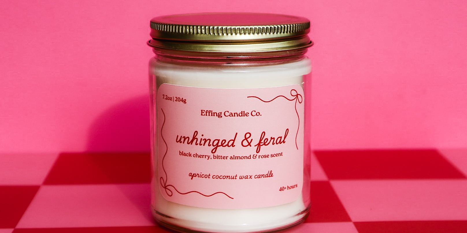 Candle jar that says Unhinged and Feral with a pink label on a red and pink checkered background