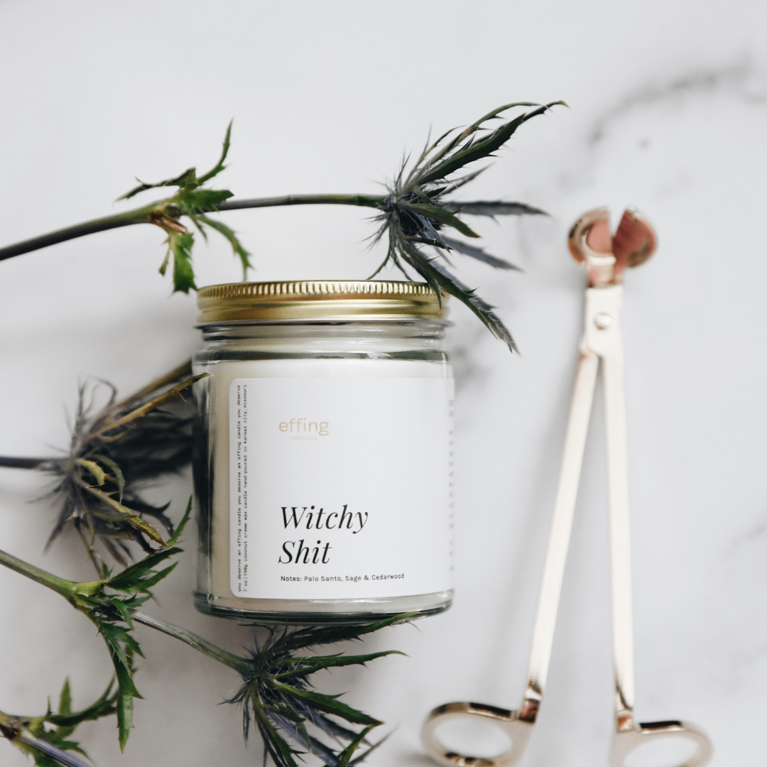 A jar candle with a wooden lid labeled 'Witchy Shit', accompanied by a metal wick trimmer and sage leaves on a marble surface.